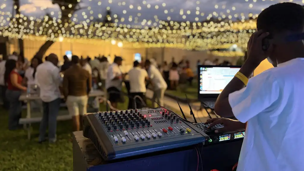 A DJ mixing at an outdoor evening event under strings of warm festoon lights, with guests gathered on the lawn at dusk